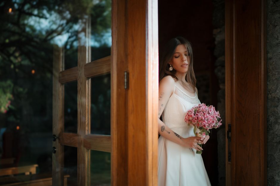 A young woman with long brown hair and tattoos on her arms is standing indoors near a wooden-framed window, holding a bouquet of fresh pink hydrangeas. She is wearing a white sleeveless dress with del
