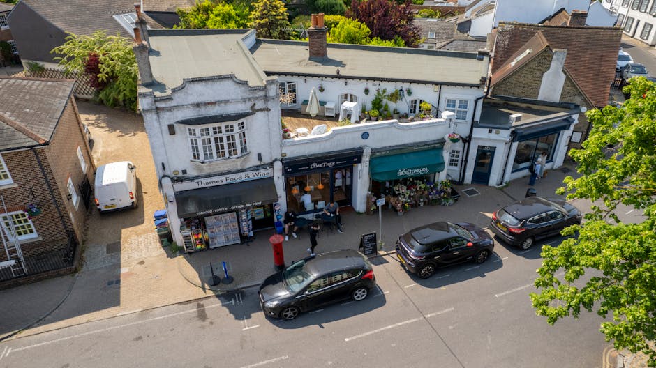 An aerial view of a quaint high street featuring a white building with a black and green awning displaying the names 'Thames Food & Wine' and a flower stand outside decorated with colorful floral arra