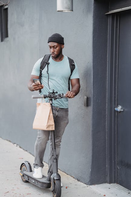 A young man with a dark complexion, wearing a black beanie, a light blue t-shirt, and grey jeans, stands outdoors against a grey wall. He holds a smartphone in his right hand while his left hand rests