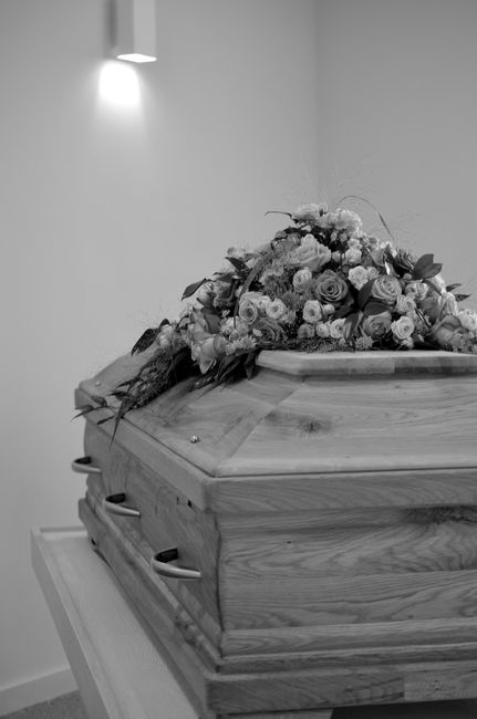 A floral arrangement of fresh, predominantly white roses and lilies with hints of pink roses, resting atop a wooden coffin in a funeral setting. The bouquet is beautifully crafted with greenery and de