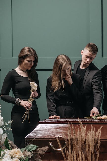 Three people at a floral arrangement event are shown over a polished wooden table with a dark green backdrop. The woman on the left, dressed in black, holds a small bouquet of white flowers with delic